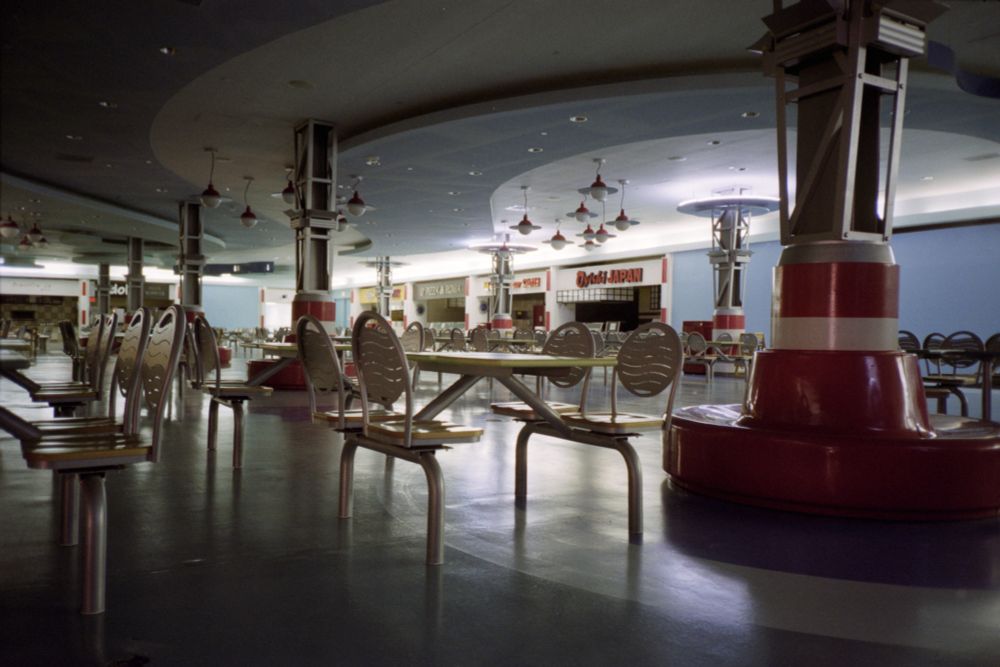 Photograph. A dimly lit, empty food court in a dead shopping mall, decorated in a Y2K-era stylized undersea theme. It is filled with empty seating and vacant eateries, in the distance.

• Olympus XA4
• Kodak Colorplus 200