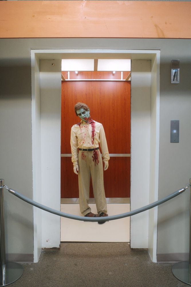 Photograph. Mannequin dressed and made-up as a zombie from the film "Dawn of the Dead" stands in the elevator formerly located in Monroeville Mall's JCPenney -- recreating a scene from the movie. White elevator doors are partially-ajar, revealing a wood and silver-trimmed cabin. Grey vinyl rope cordons off the display.