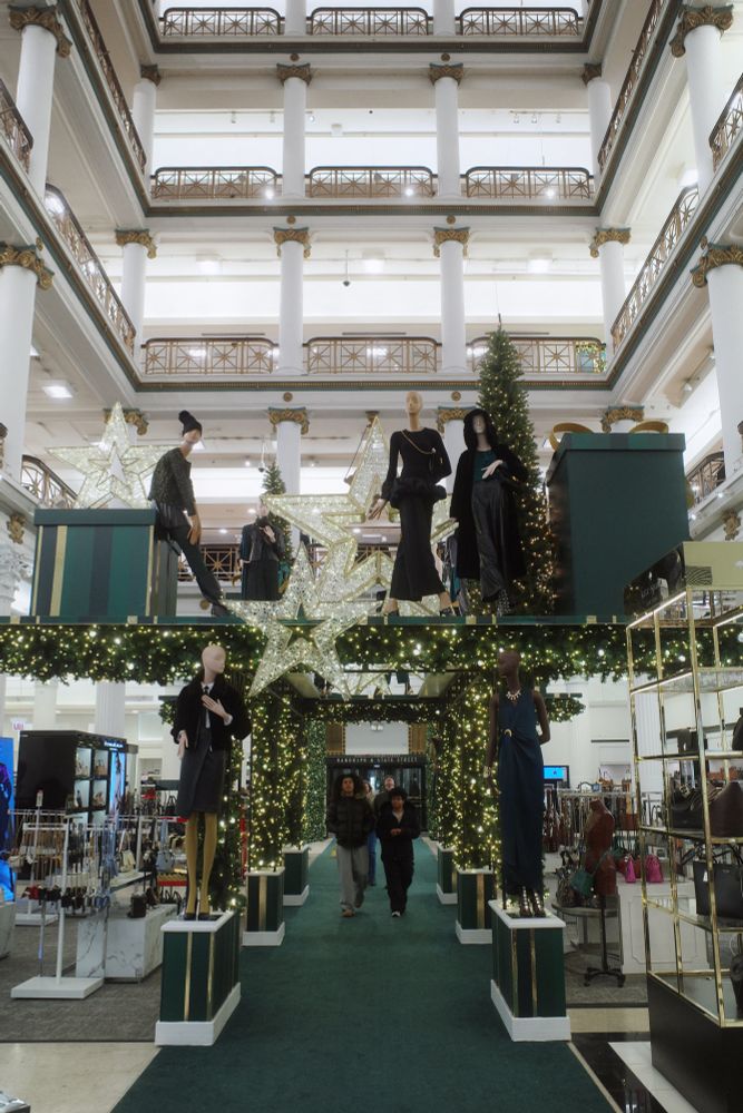 Photograph. A large department store atrium with towering neoclassical columns rising over the sales floor. Shoppers pass under a Holiday display of green gift boxes and stylish mannequins. The carpet is green opposed to Macy's usual red - more nods to the Marshall Field's aesthetic.
