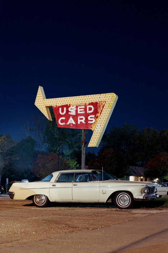 Night photograph of a mid-century Chrysler Imperial sedan parked under a weathered, period-correct dealership sign that reads "USED CARS"