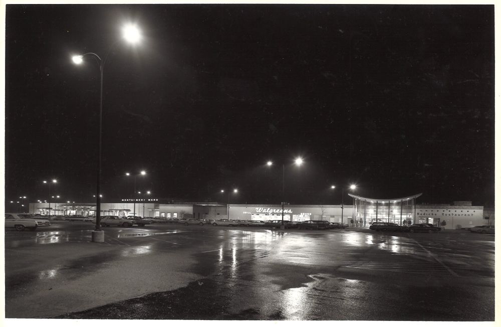 Photograph. An enclosed shopping center at night as viewed from the end of a wet parking lot. Stores visible include Montgomery Ward, Walgreen's and Nedick's Restaurant. A curved entryway serves as the mall's main entrance.