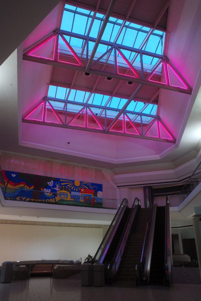 Photograph. A dark atrium in a bilevel shopping center. A vacant department store anchors the end, no retailers are open in this frame. A pair of nonworking escalators with a central staircase to the right. An empty conversation pit, below. A large skylight overhead features oversized support trusses, accented by pink neon inserts. Blue hour outside.