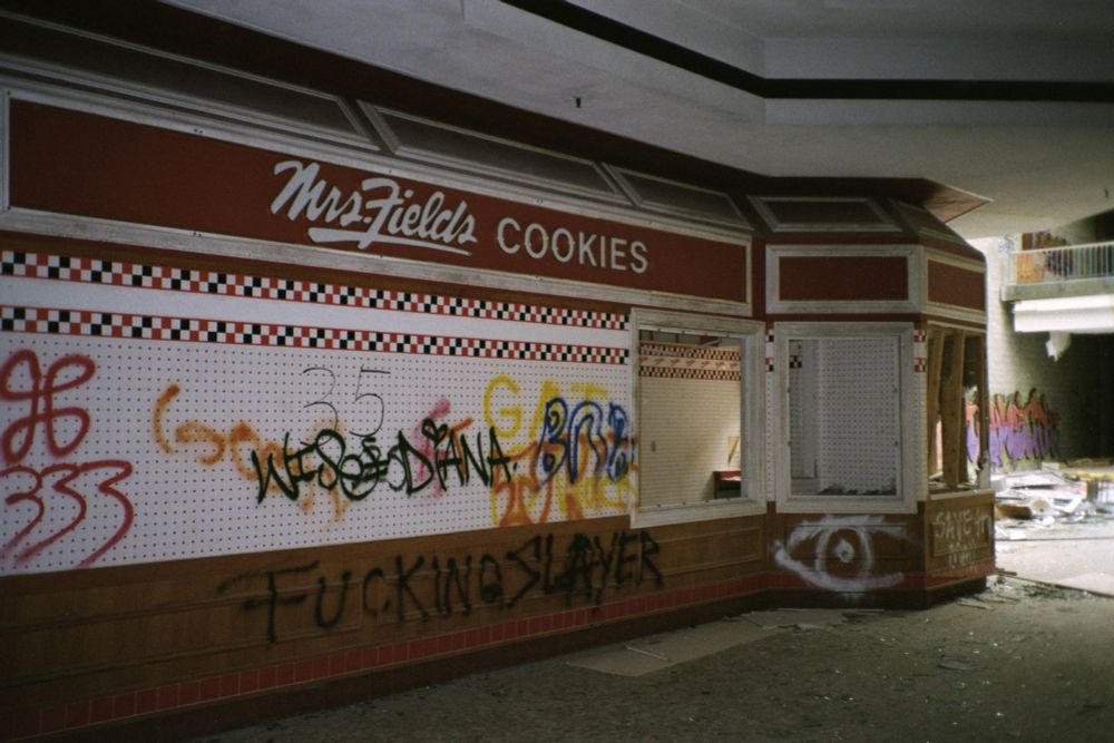 Lo-fi film photograph of a Mrs. Field's Cookies store in an abandoned mall. The facade is covered in graffiti. Natural light pours in through the atrium, in the background.
