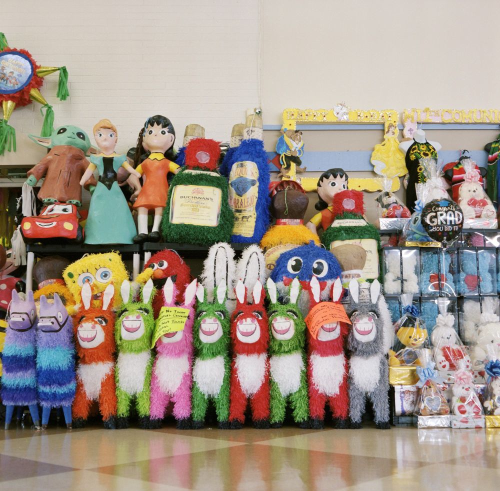 Photograph. A group of pinatas in both traditional style and IP's on display in an enclosed mall concourse. Large frames and gift baskets are also seen to the right.