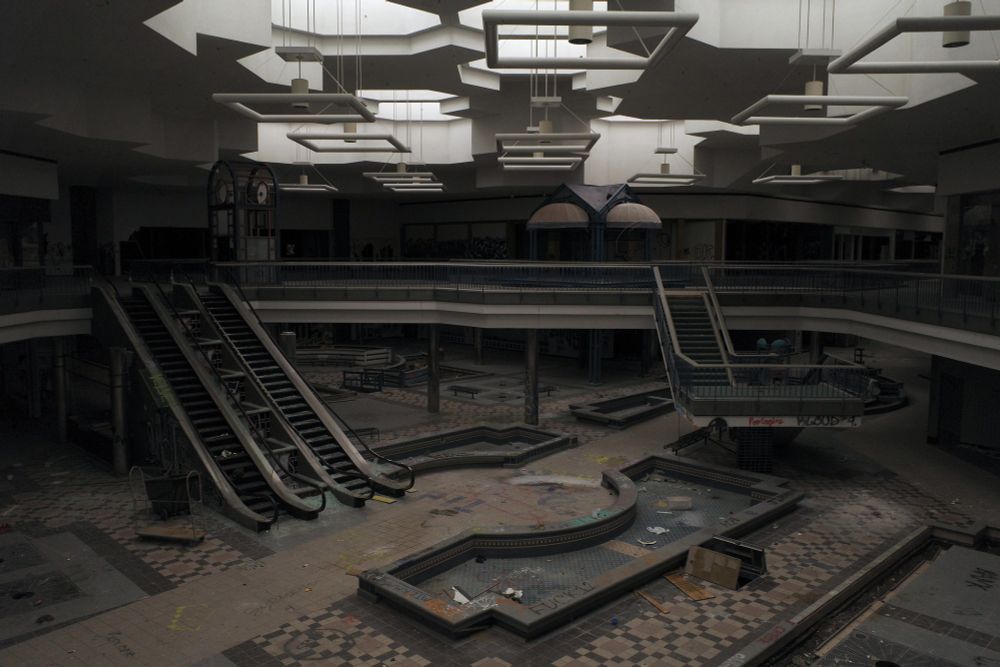 Photograph of an abandoned two-level shopping mall's center court. The lighting is dim, it is a 10-minute long exposure illuminated only by moonlight coming through the skylights. Debris and graffiti are visible throughout the scene, amid escalators, fountain pits and staircases.