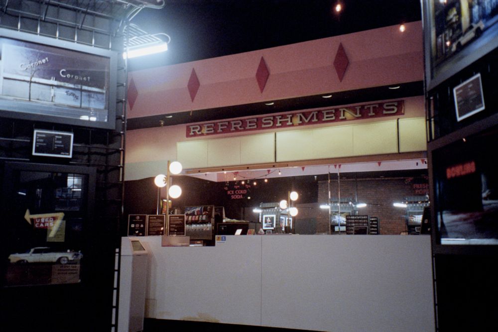 Photograph. A mid-century cinema lobby with a concession stand and art gallery. Photography displays flank both sides of frame.