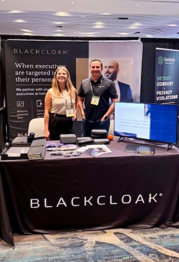 Two people standing at the BlackCloak exhibition booth, smiling at a convention. The booth features informative displays and promotional materials.