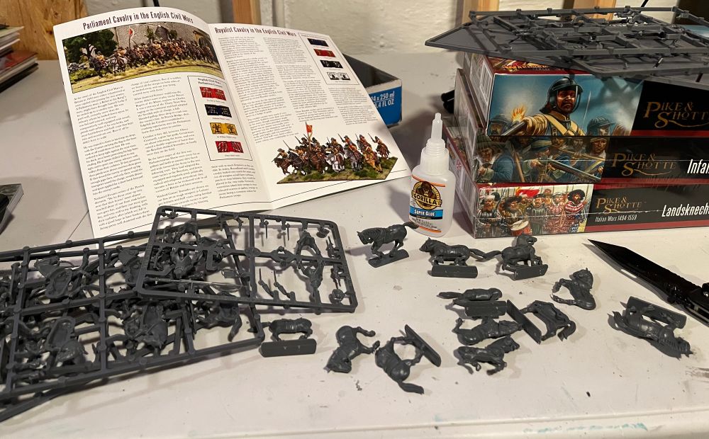 Photo of plastic miniature horse Calvary being assembled on a table. There’s a pamphlet propped up in the back and 3 boxes of miniature English Civil War soldiers on the right.