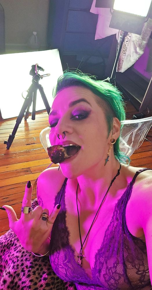 A selfie of Gemma Rae wearing a sheer purple lace top and heavy eye makeup, they're leaning on a leopard print bed spread, grinning at the camera around a chunk of cake in their mouth and throwing the horns. They're limned in neon pink light, and behind them on the wooden floor is a camera tripod, a soft box video light, and another tripod with a video light on top. 