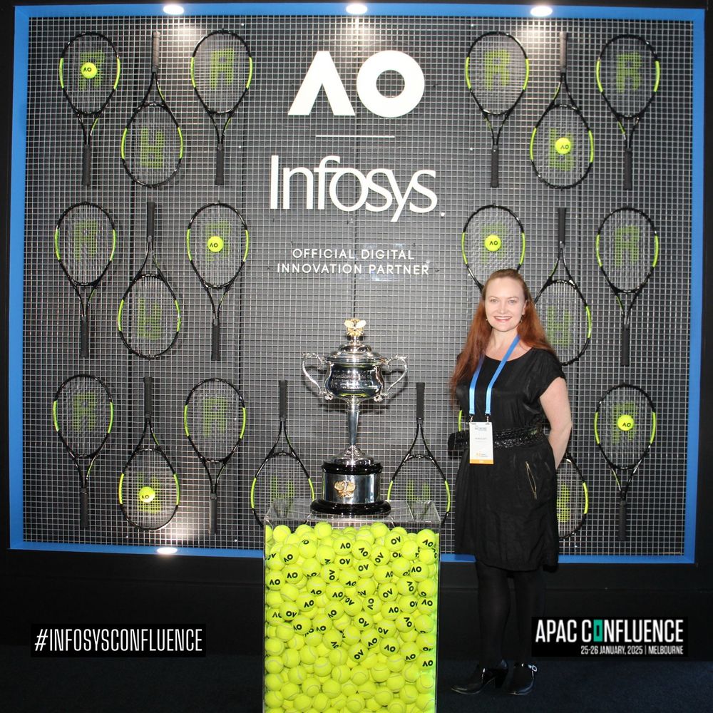 A redhead woman stands in front of a branded tennis racket wall, next to the Australian Open Tennis womens’ championship trophy. 