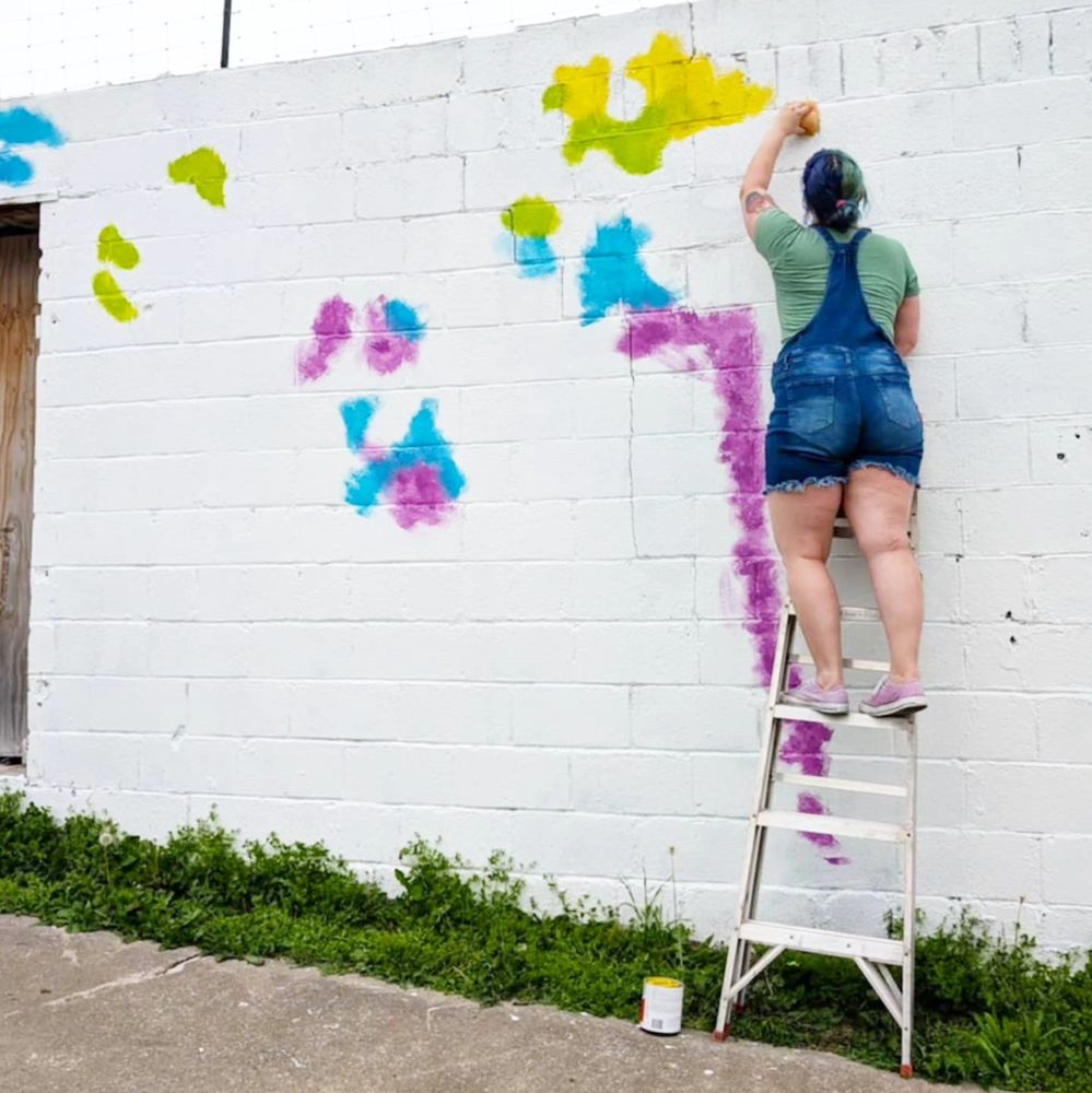 A woman with blue hair stands on a ladder, painting colorful splatters on a white wall. 