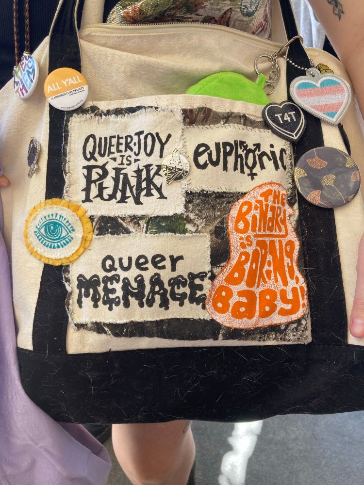 a photo of a black and white tote bag with several patches and pins sewn/stuck to it. two of Cody’s previously shown patches are there, as well as a third that says “euphoric” with a trans symbol in the center as the letter “o”. another patch (not made by Cody) says in orange “the binary is boring, baby”