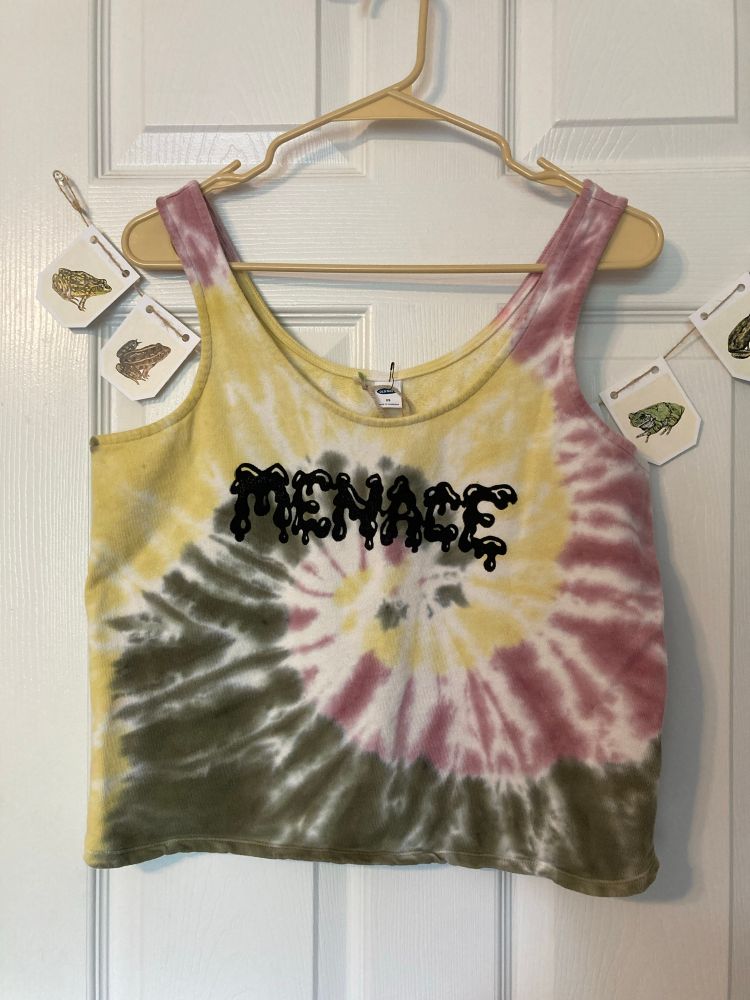 a photo of a tie-dyed french terry tank top crop in non-binary colors. the middle says “MENACE” printed in black ink. it has a drippy liquid font. 