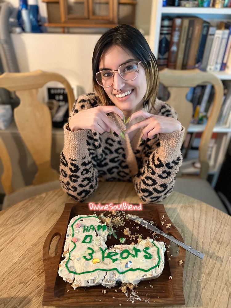 Nemea sitting at a wooden table with a wooden tray and a partially eaten homemade cake. 3 years can be seen on the cake in geek icing. There is a silver knife on the tray. Nemea is smiling holding their fingers in a heart shape. Smiling at the camera wearing glasses, nose piercings, fake nails, makeup and a cheetah print cardigan. The DivineSoulBlend watermark is in the center. There are wooden chairs, a wooden bookcase filled and a shelf with items behind Nemea. 