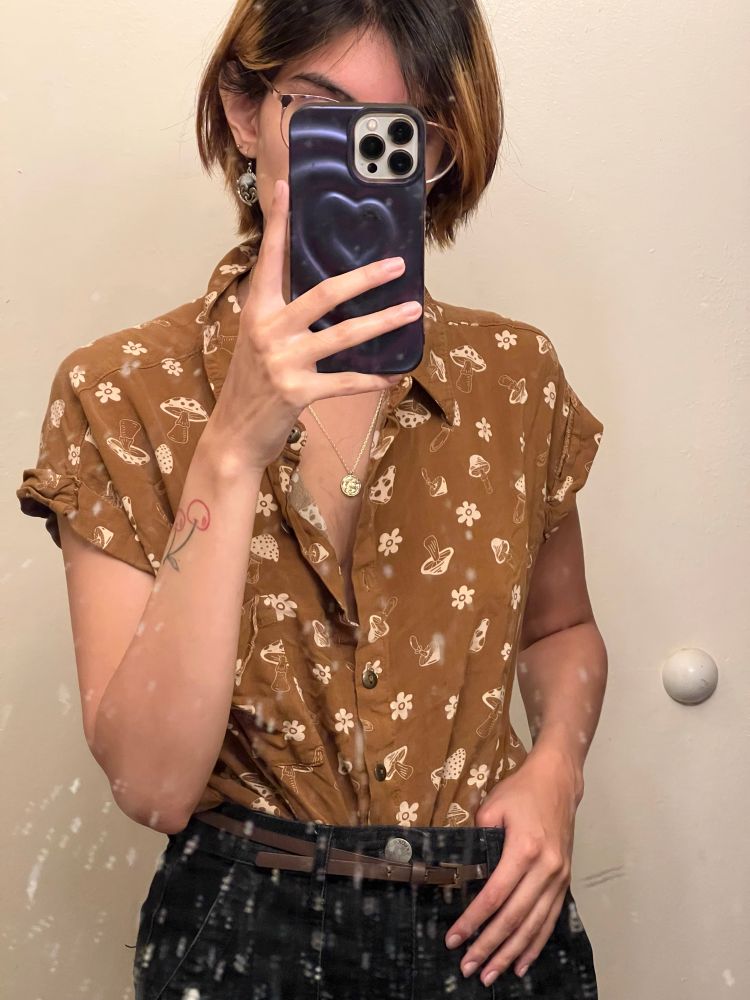 Nemea wearing a brown top with yellow different types of mushrooms. They have one hand holding the front of their jeans and brown belt. They have a gold chain necklace with a gold pendant. Their cherry tattoo is visible as the other hand is holding up their phone which is covering most of their face. They have mushroom earrings on, glasses and their partially dyed head. 