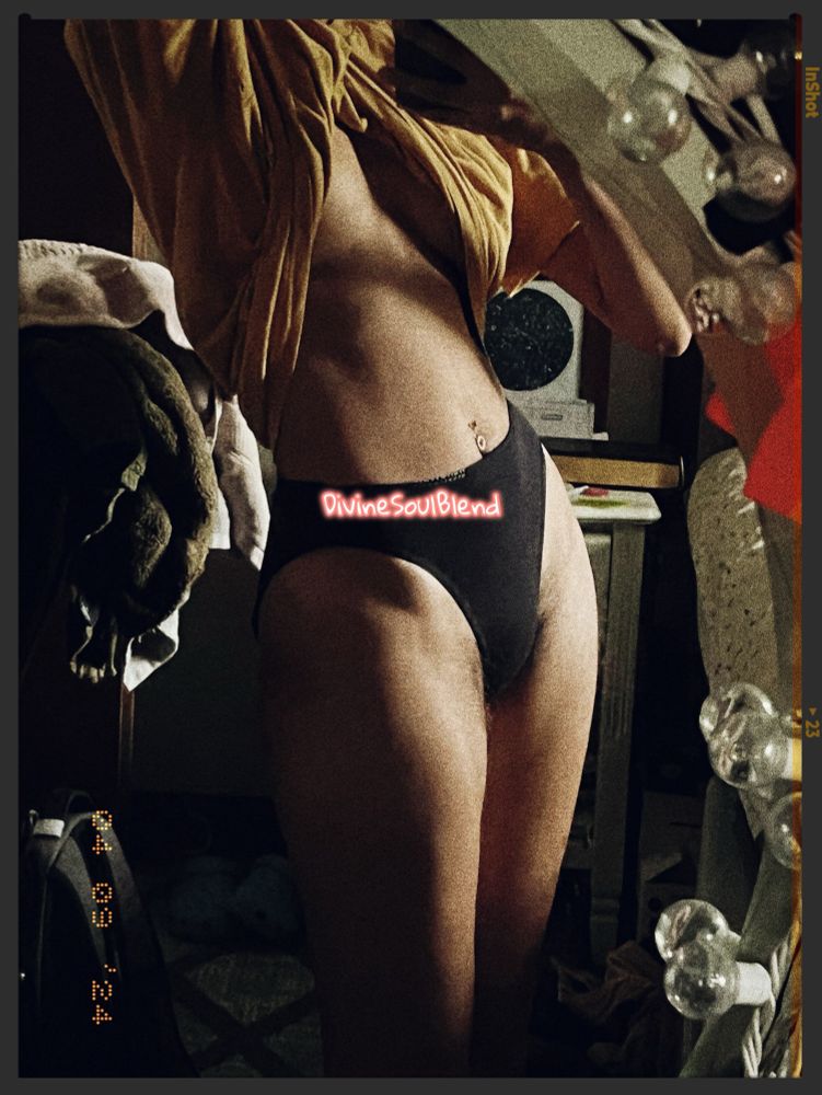 A mirror photo of Nemea which focuses on their chest to feyr panties. The frame shows from feyr legs to their neck. Nemea is holding up some of their yellow top. Feyr wearing black panties and no bra. The light is hitting part of their chest and feyr thighs. The mirror has bounced lights. There is a chair with some towels folded over. They have a belly piercing. The DivineSoulBlend watermark is visible