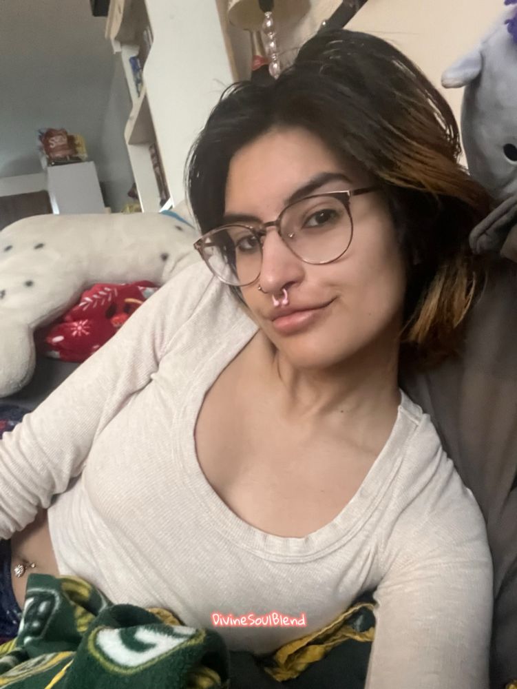 Nemea laying on a grey couch with a Green Bay packer blanket. They’re smiling at the camera with a head tilt. They have glasses on and nose piercings. They are wearing a cream top. There is a white cabinet behind them and a big stuffed animal. The Divinesoulblend watermark is towards the center bottom of the pic 