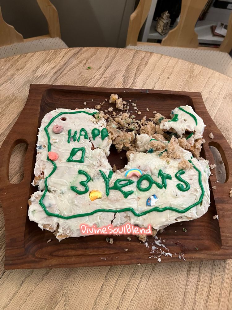 On a wooden table there is a darker brown tray with a partially intact cake. The cake has been partially eaten and destroyed. In green “3 years” can be read on the cake. There is towards the outer edge a chair and part of a bookshelf with a wooden robot. The pink DivineSoulBlend watermark is on the center of the cake 