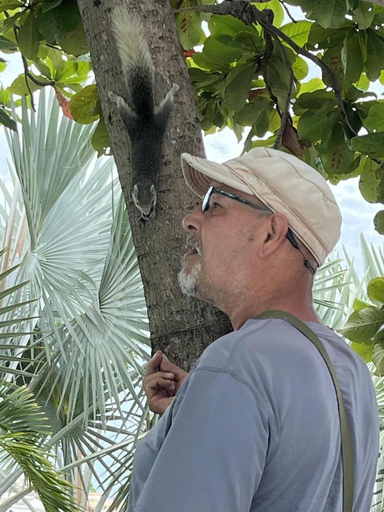 Old guy with a cap talking to a squirrel, because it's there and is busy eating, so of course old guy must be a pain and share his day, knowing full well 🐿️ has seen the next nut being proposed and so decides it's worth putting up with his big nose nearby and that weird breath.. from it's 🤔 snout