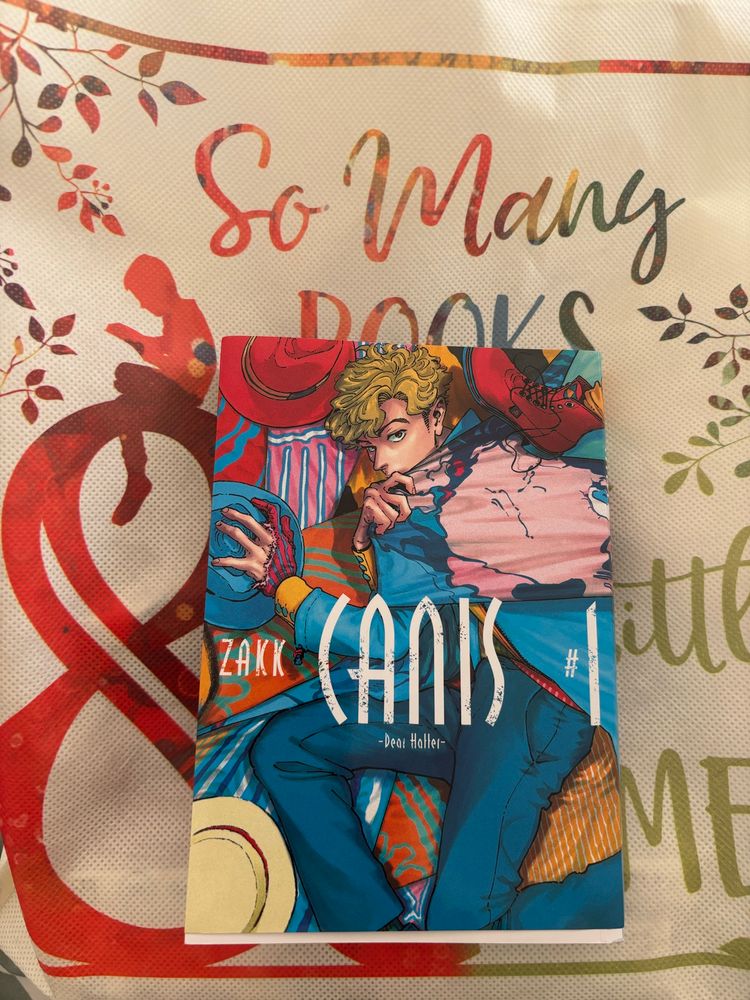 A cream colored B&N bag with orange lettering in the back ground and Zakk’s Canis-Dear Hatter- #1 is on top of it with the main character Kutsuna surrounded by various hats and a suspicious blue fabric that looks like it was Ryou’s face, blue eyes, and black haired being pulled across Kutsuna’s shoulder. 