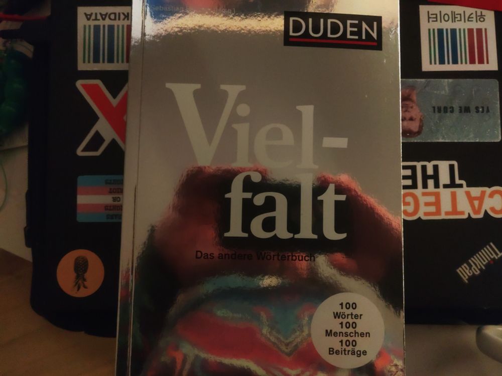 Der Vielfalt-Duden auf einem bestickerten Laptop, unter anderem sind ein Ananas-Sticker, ein xmonad-Sticker, ein Category-Theory-Sticker, ein Yes-we-curl-Sticker und zwei Wikidata-Sticker zu sehen.