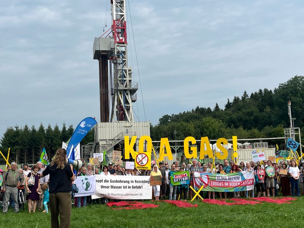 Das Foto zeigt den Koa Gas Protest in Reichling direkt am Bohrturm. Darauf zu sehen sind Demonstrierende mit vielen Schildern, gelben Protestkreuzen und dem Slogan „Koa Gas“.