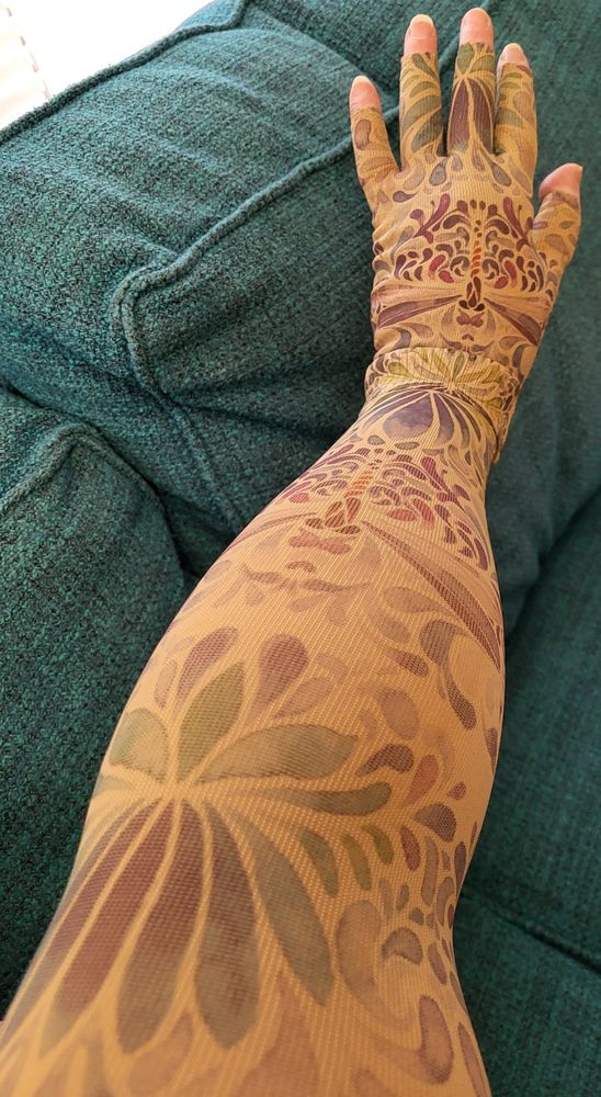 My outstretched arm and hand, clad in compression sleeve and glove that have a beige background with a pattern of dragonflies, leaves, and flowers in subtle shades of blue, green, and purple. 