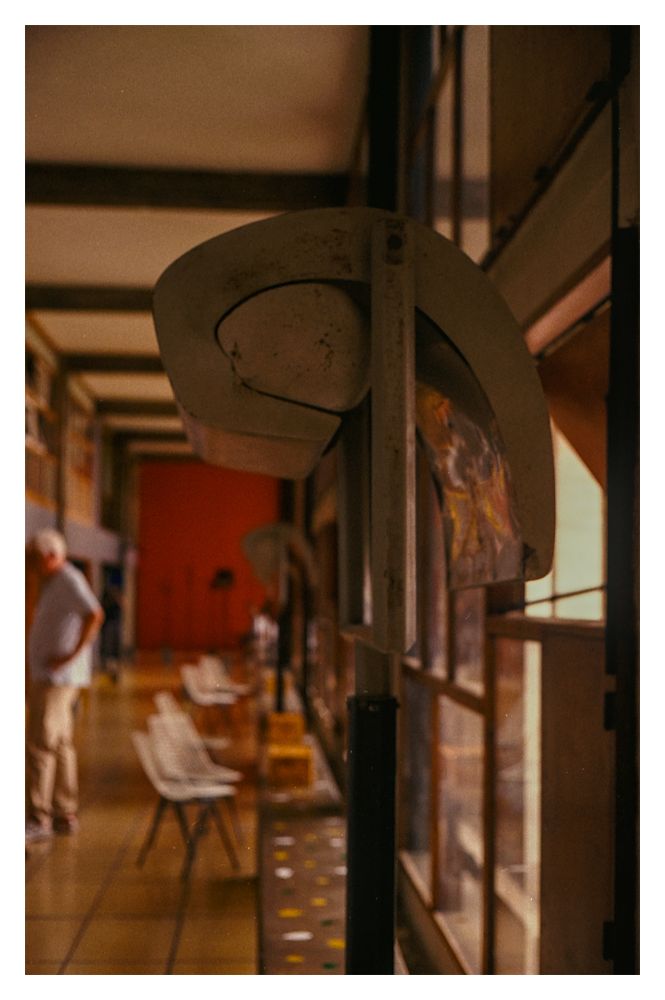 Color photograph taken in Le Corbusier's Cité Radieuse in Marseille. In the foreground, a close-up of a curved metal bracket mounted on the wall is clearly visible. Behind it, a long interior corridor stretches into the distance, slightly out of focus. The corridor has large windows on the right side through which warm light enters, a tiled floor with small colored dots, and a row of white chairs along the wall. Further back in the hallway stands a person who is partially out of focus. The overall color tone is warm, with brown and orange tones and soft shadows.


Translated with DeepL.com (free version)