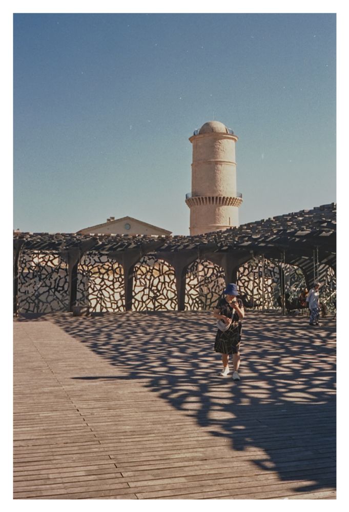 Color photograph taken on the terrace of the Mucem - Musée des civilisations de l’Europe et de la Méditerranée. The terrace is enclosed by a net-like concrete structure that casts a playful shadow on the wooden floor. A woman walks toward the lens with her gaze lowered and a sun hat covering her face. The Tour du Fanal can be seen in the background. This tower, built in 1644, served as a guide tower at the harbor and guided ships safely in. The tower and the mesh concrete are surrounded by a blue, cloudless sky.

Translated with DeepL.com (free version)