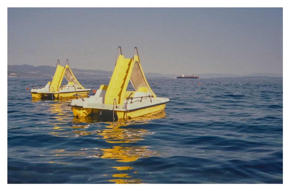 Color photograph of two yellow pedal boats with slides floating in the blue water off the beach at Opatija. A container ship can be seen further out at sea in the background.