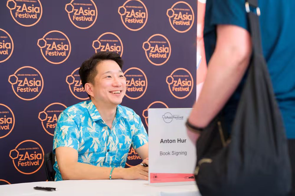 Anton in a blue shirt sitting against an OzAsia Festival backdrop at a book signing