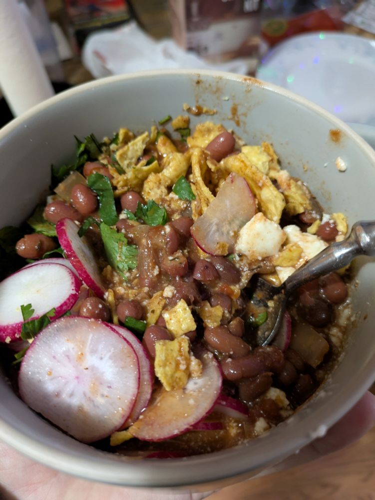A bowl of thick bean soup with queso fresco, radish, cilantro and crushed tortilla chips with beans, tomatoes, onions, garlic and spices, mixed up a bit!