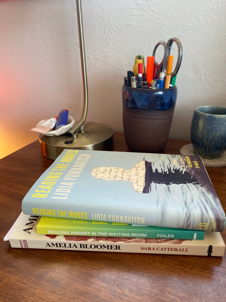 A pile of three books: Reading the Waves, Reading Arendt in the Waiting Room, and Amelia Bloomer. In the background: a brown pen cup, a gold lamp, a blue water cup. 