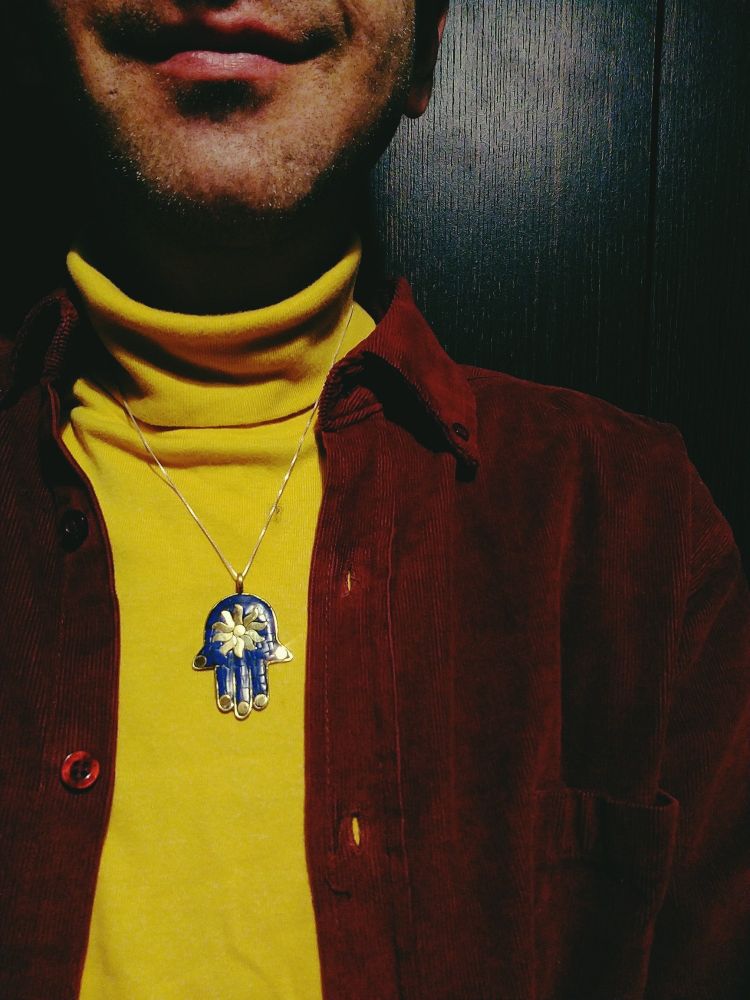 Un sorriso stupido, una barba cortissima, un lupetto giallo, una camicia di velluto a coste rossa e una mano di fatima blu con un sole dorato al centro