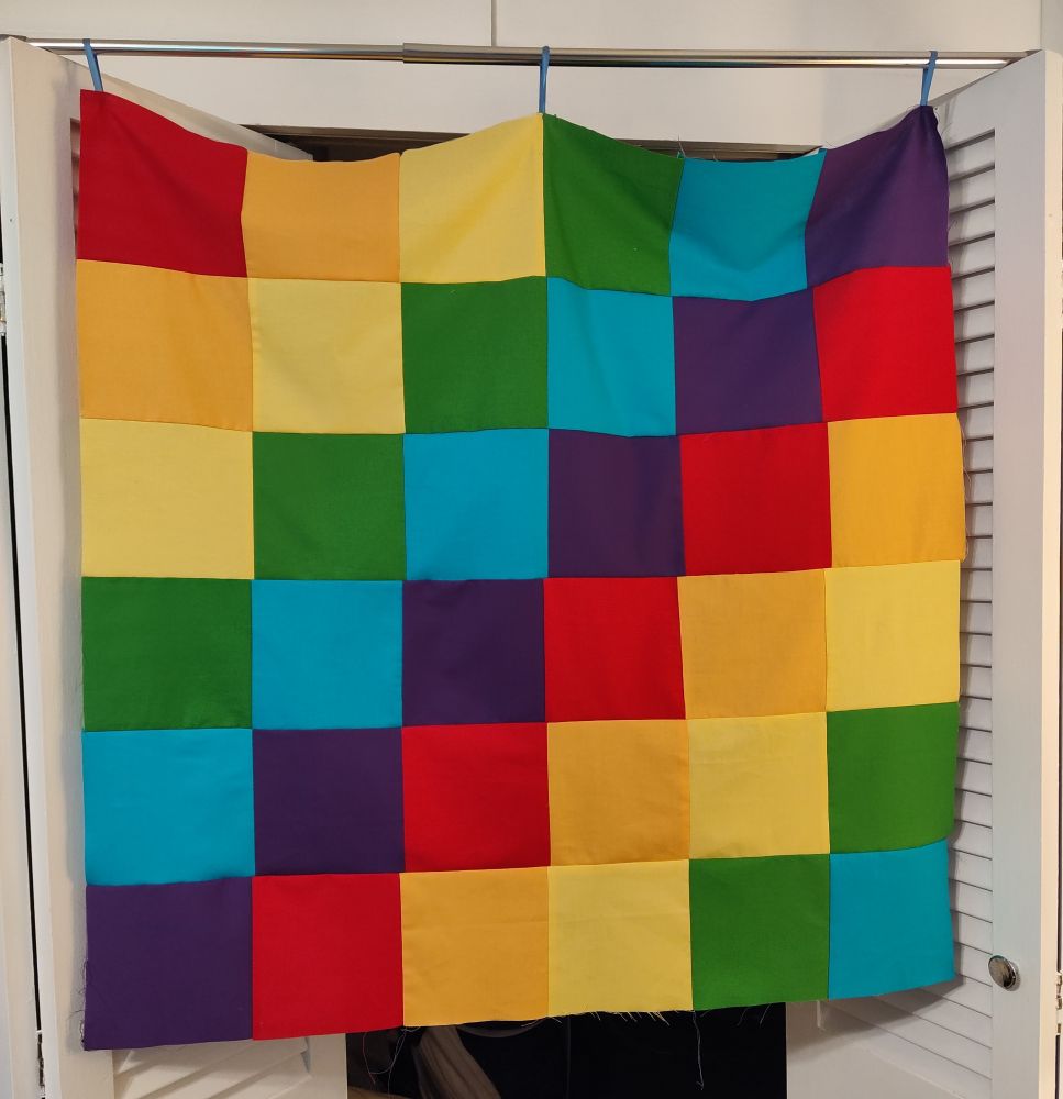A baby-sized quilt top composed of solid squares in rainbow colors hangs from a curtain rod.