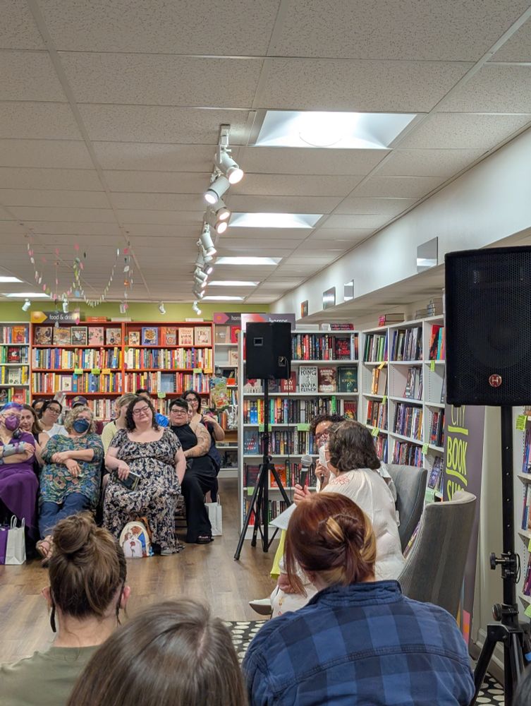 The view from my seat, showing Mia Sosa interviewing Olivia Dade in the downstairs area of East City Bookshop, centered between two speakers, in front of many bookshelves