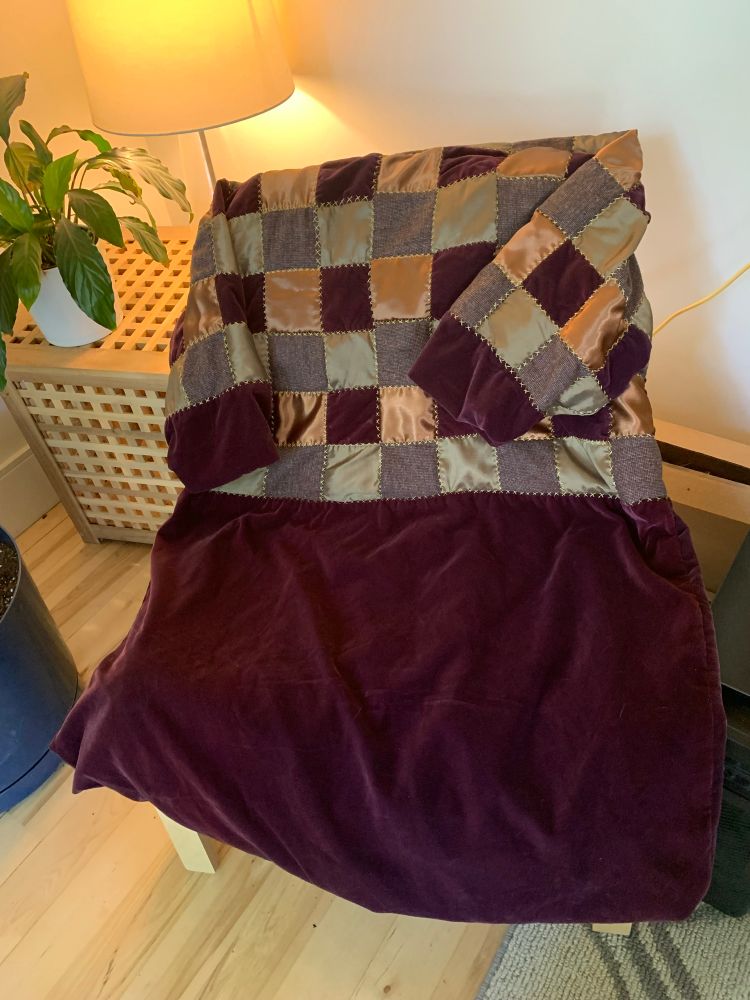 The back of a purple velvet, brown satin, green polyester and brown wool patchwork knee-length jacket with a shawl collar and brown and gold buttons resting on a chair with a plant and lamp in the background 
