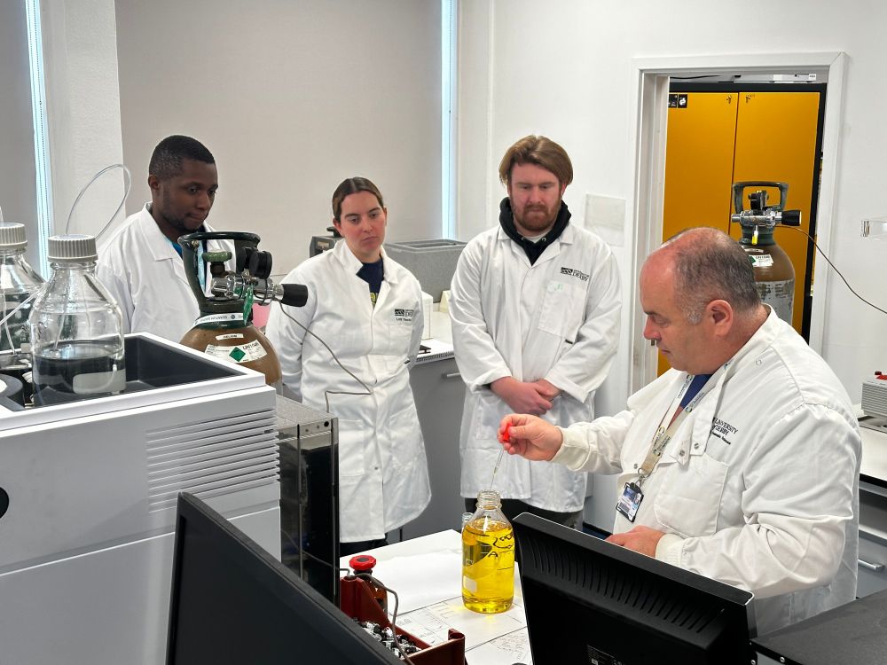 Laboratory technician at the University of Derby extracting a fluid sample from a glass container being watched by students.