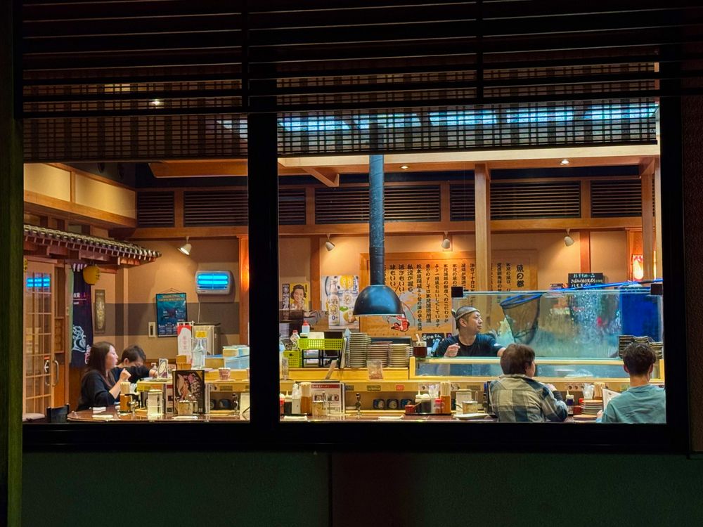 The view from outside a sushi restaurant.  It is dark outside and we see a colorful scene through the window.  A chef stands behind the counter next to a large fish tank and four diners are arrayed around him at the bar.  The wooden walls are covered in descriptions of menu items.