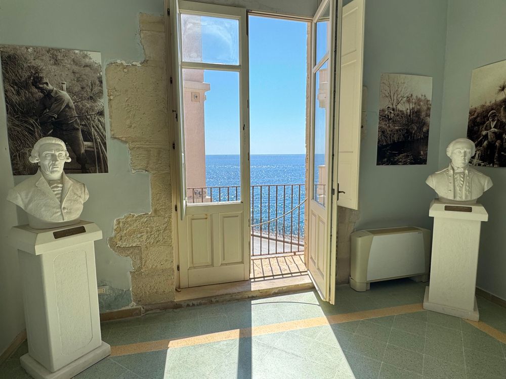 A room in the museum.  Two statuary busts are in front of an open window framing a bright blue sea.

