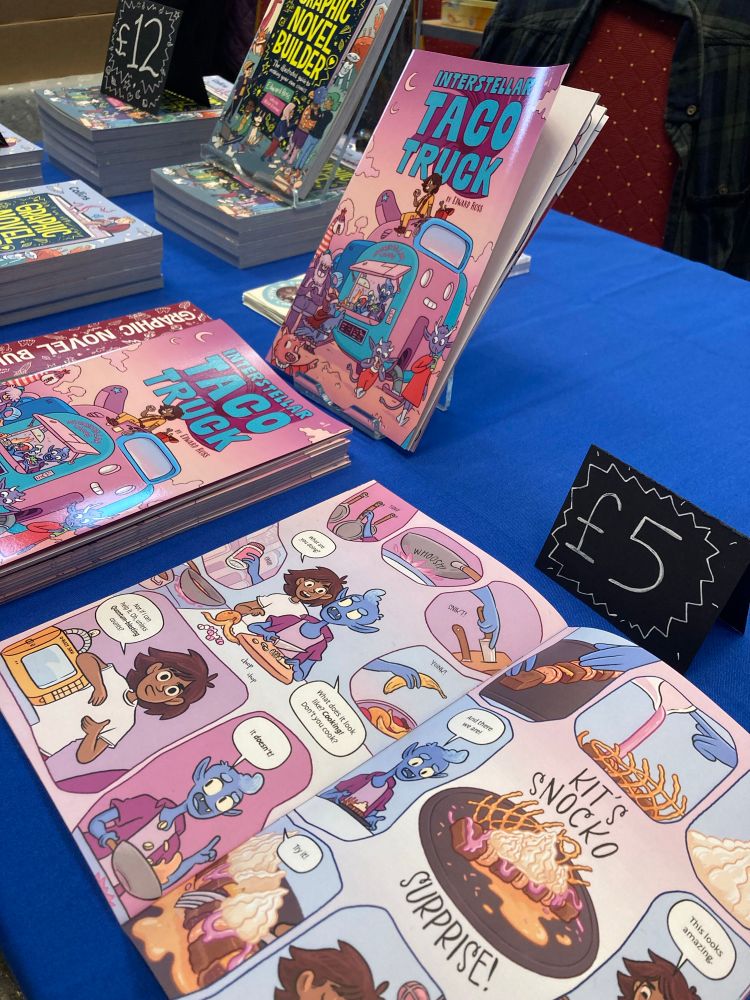 Convention table featuring copies of my mini comic Interstellar Taco Truck. A pink, blue and purple colour scheme. We see human and alien kids cooking a delicious looking pudding!