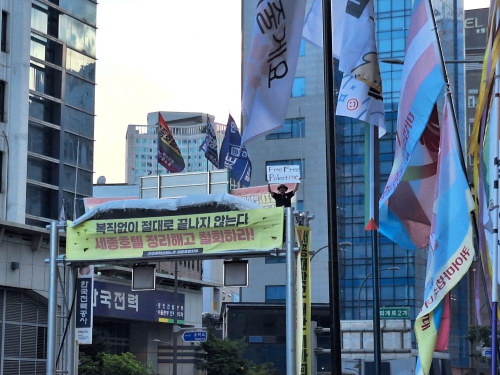 세종호텔 앞. 고공농성 중인 사람이 행진해온 사람들에게 "Free Free Palestine" 피켓을 들어올려 보여주고 있는 사진.