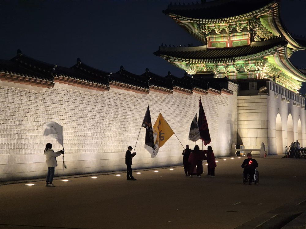 밤의 광화문 돌담 옆에서 사람들이 깃발을 들고 있다.