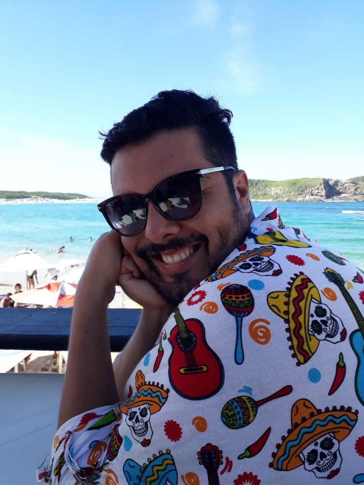 My very handsome husband, Rafael, posing for a picture at the beach. He is sitting at a table with the beach behind him, wrapped in a cute and funny beach towel (white with a Mexican motif), wearing dark sunglasses and a cute smile.