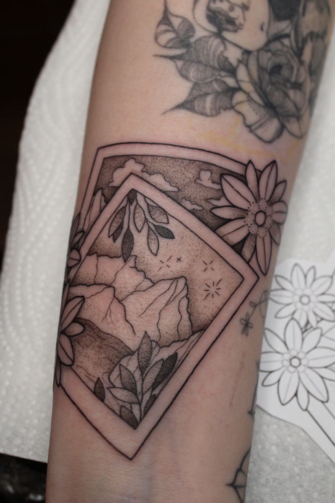 A tattoo of two Polaroids, the top one showing mountains and a lake. Surrounding the Polaroids are edelweiss flowers