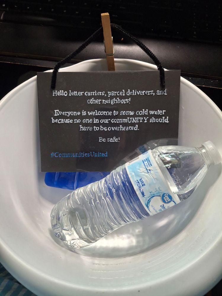 White plastic bowl with a bottle of water and a freeze pack in it, with a small black sign saying "Hello letter carriers, parcel deliverers, and other neighbors! Everyone is welcome to some cold water because no one in our commUNITY should have to be overheated. Be safe!" The hashtag #CommunitiesUnited is also on the sign.