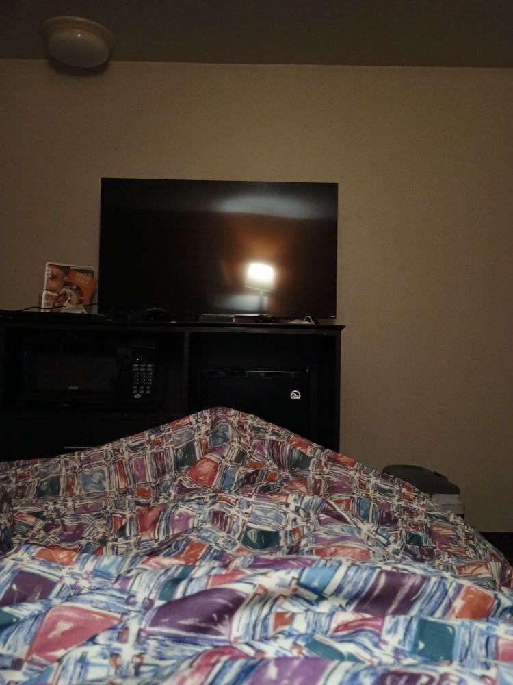 Point of view photo from lying in a bed. The blanket boasts a hideous pattern of differing colored squares that look like they were drawn on the fabric with a marker. At the foot of the bed is a black cabinet with a microwave and refrigerator below and a large rectangular television looming above. The walls are a sickening shade of off-white tinted by a warm light that can be seen reflected in the otherwise dead visage of the TV. There is a strange bulbous light, dark and cold, watching from the ceiling above. You can almost smell the acrid cleaning chemicals and discount carpet perfume leaking off the image. This is a hotel room, a cheap hotel room at that.