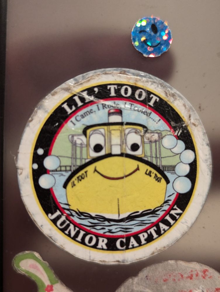 A happy little yellow cartoon boat sails through a harbor. One ring of text around it reads "Lil' Toot Junior Captain", and a smaller inscription reads, "I Came, I Rode, I Tooted....!"