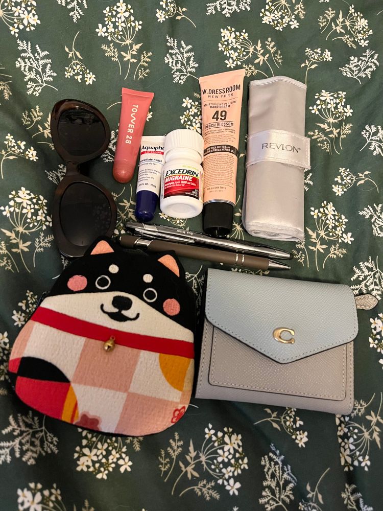 The contents of VTuber Nova Eos's purse. From left to right, top to bottom, the items are as follows: a pair of brown cateye sunglasses, a Tower28 lip treatment, Aquaphor lipbalm, a small container of medicine, W. Dressroom Peach Blossom hand cream, a silver nail kit, two pens, a small pouch shaped like a black shibu with a red collar and bell, and a small, gray and baby blue Coach wallet. The items are laid out on a dark green bedspread patterned with white, cream, and beige flowers.