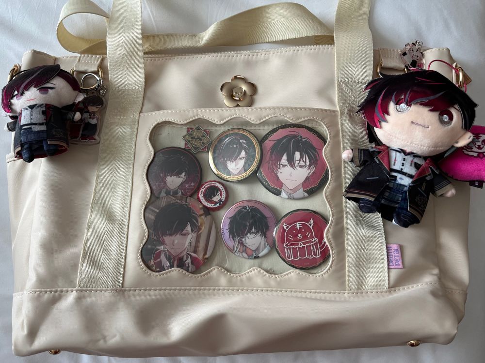 A cream itabag tote laying on a white duvet cover. The itabag features various keychains, plushies, pins, and charms of VTuber Ver Vermillion from NIJISANJI EN's 7th wave XSOLEIL.